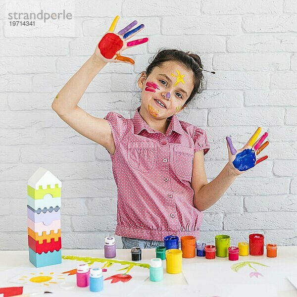 Nettes kleines Mädchen zeigt gemalte Hände stehend vor Tisch mit bunten Farben