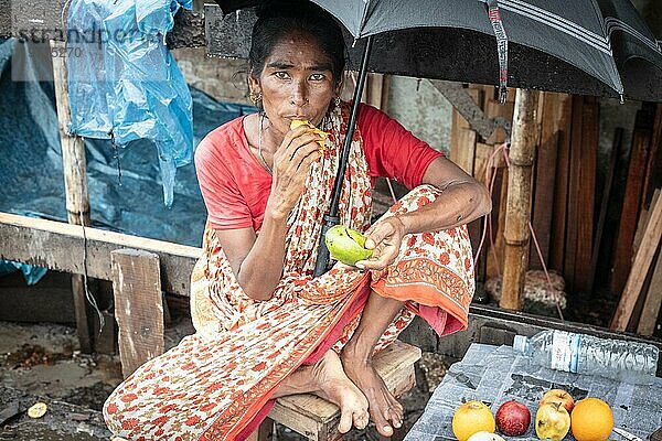 Frau isst eine Mango vor ihrem Haus  sie schützt sich mit einem Regenschirm vor einem Monsunschauer  Tejgaon Slum Area  Dhaka  Bangladesch  Asien