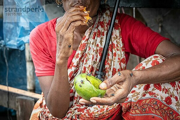 Frau isst eine Mango vor ihrem Haus  sie schützt sich mit einem Regenschirm vor einem Monsunschauer  Tejgaon Slum Area  Dhaka  Bangladesch  Asien