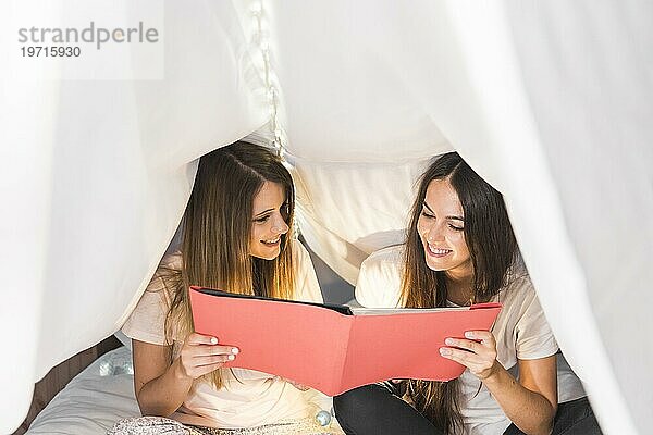 Zwei glückliche Freundinnen schauen Album sitzend Bett weißen Vorhang
