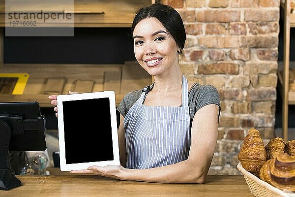 Lächelndes Porträt einer jungen Bäckerin  die ein digitales Tablet in der Hand hält  das an einer Bäckertheke steht