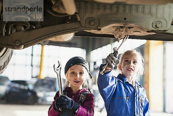 Zwei kleine Mädchen in Overalls reparieren ein Auto mit Schraubenschlüsseln