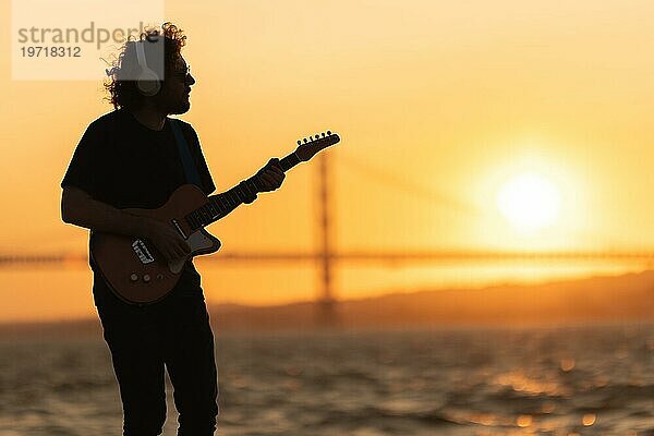 Romantischer Mann mit kabellosen Kopfhörern steht am Wasser und spielt Gitarre bei orangefarbenem Sonnenuntergang. Mittlere Aufnahme