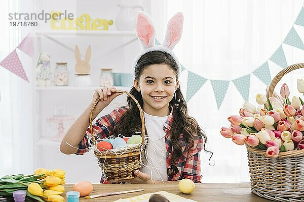 Glückliches Mädchen mit Hasenohren hält Korb mit bunten Ostereiern