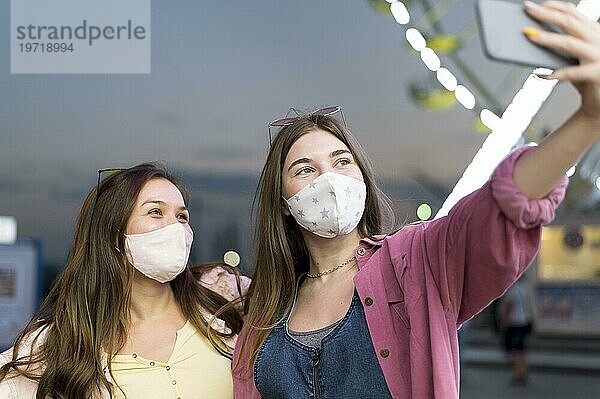 Weibliche Freunde mit Masken nehmen Selfie Vergnügungspark