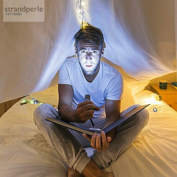 Mann sitzend Vorhang Bett hält Album beleuchtet sein Gesicht mit Taschenlampe