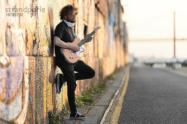Romantischer Hipster mit Sonnenbrille an der Wand stehend und E Gitarre spielend bei frühem Sonnenuntergang. Mittlere Aufnahme