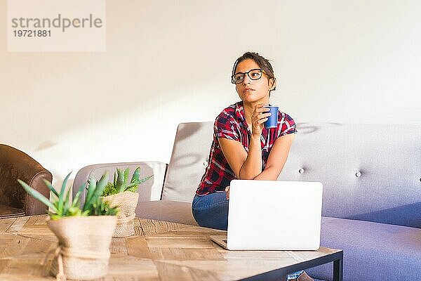 Junge Frau an einem Arbeitstag zu Hause im Wohnzimmer sitzend mit einer Tasse Kaffee