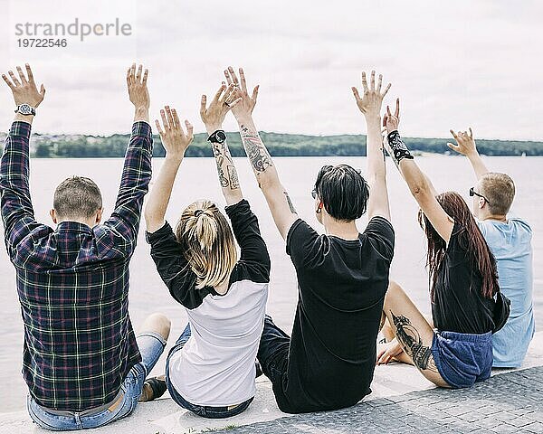 Rückansicht Freunde  die in der Nähe des Sees sitzen und ihre Hände heben