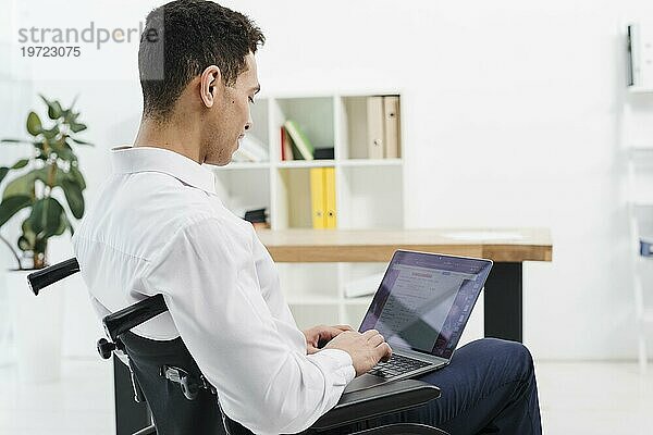 Seitenansicht behinderter junger Mann im Rollstuhl sitzend mit Laptop Büro