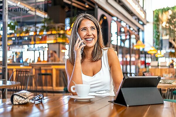 Abgelenkte Geschäftsfrau  die in einer Cafeteria mit einem digitalen Tablet und einer Kaffeetasse sitzt und telefoniert