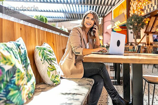 Lächelnde Frau  die einen Laptop benutzt und mit dem Handy in einer Cafeteria spricht