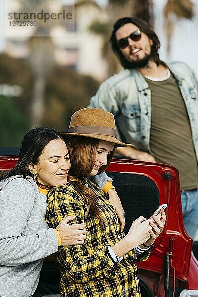 Fröhliche Frauen umarmen Dame mit Smartphone in der Nähe von Auto Kofferraum Mann lehnt sich aus dem Auto