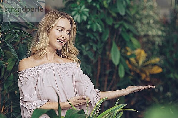 Lächelnde blonde junge Frau mit offener Handfläche in der Nähe einer Pflanze stehend