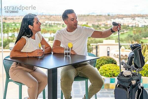 Reicher Mann hält einen Golfschläger in der Hand und unterhält sich mit seinem Paar  während er auf einer Terrasse Cocktails trinkt