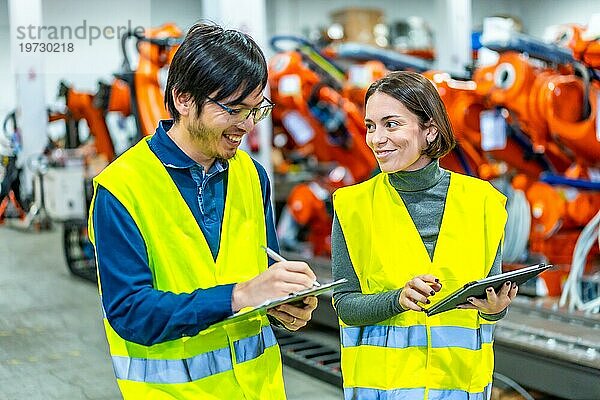 Multikulturelle männliche und weibliche Ingenieure lächeln  während sie das Fließband einer Fabrik für Roboterarme kontrollieren