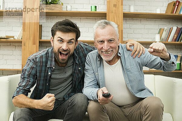 Älterer glücklicher Mann mit Fernbedienung junger weinender Mann vor dem Fernsehsofa