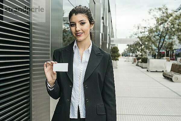 Glückliche junge Geschäftsfrau steht vor einem Bürogebäude und zeigt ihre Visitenkarte