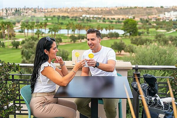 Lässiges  reiches Paar  das auf einer Terrasse mit Blick auf einen Golfplatz mit Cocktails anstößt