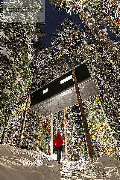 Tourist bewundert die erhöhte Hotelzimmerkapsel in Form zwischen verschneiten Bäumen im borealen Wald  Schwedisch-Lappland  Harads  Schweden  Skandinavien  Europa