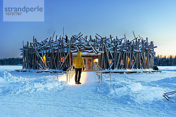 Person steht auf der Brücke  die das Hauptgebäude des beleuchteten Arctic Bath Hotels aus Baumstämmen verbindet  Abenddämmerung  Harads  Schwedisch-Lappland  Norrbotten  Schweden  Skandinavien  Europa