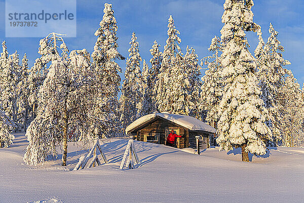 Tourist steht in der frühen Morgensonne vor einem isolierten Chalet im verschneiten Wald  Norrbotten  Schwedisch-Lappland  Schweden  Skandinavien  Europa
