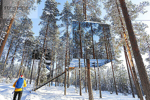 Blick auf einen Touristen  der den würfelförmigen Raum mit Spiegelwand in einem mit Eis und Schnee bedeckten borealen Wald bewundert  Tree Hotel  Harads  Lappland  Schweden  Skandinavien  Europa