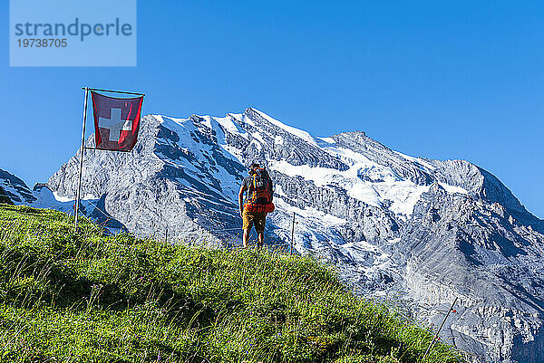 Rückansicht eines Mannes mit Rucksack beim Wandern durch die Schweizer Alpen  vorbei an einer wehenden Schweizer Flagge  Oeschinensee  Kandersteg  Kanton Bern  Schweiz  Europa