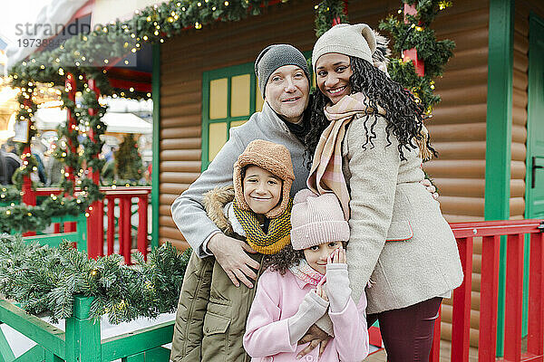 Glückliche Familie steht zusammen vor einem Holzhaus auf dem Weihnachtsmarkt