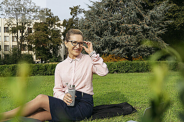 Smiling businesswoman adjusting eyeglasses sitting on grass in park