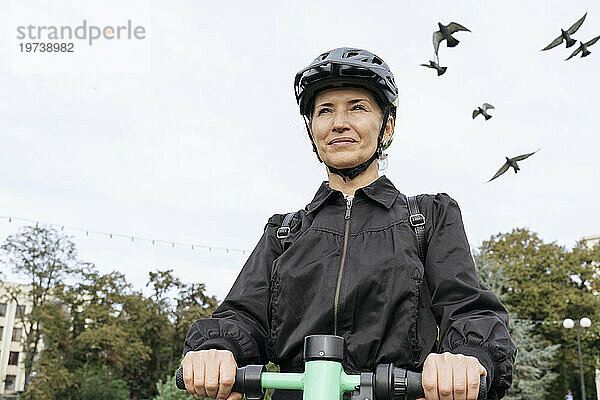 Glückliche Frau mit Helm und Elektroroller im Park unter freiem Himmel