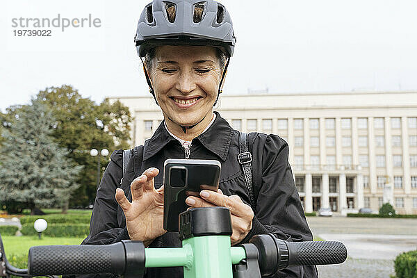 Lächelnde Frau steht mit Elektroroller und benutzt Smartphone im Park