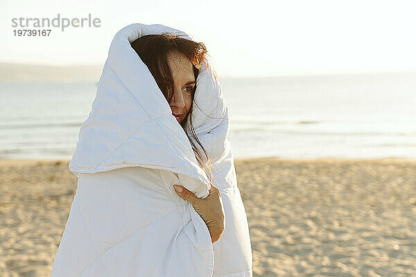 Lächelnde Frau  in eine Decke gehüllt  steht am Strand