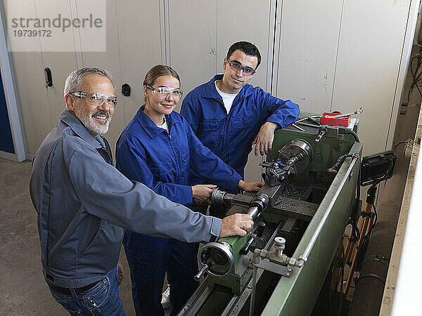 Smiling instructor standing with apprentices near lathe machine at workshop
