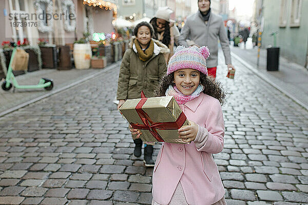 Lächelndes Mädchen hält Geschenkbox in der Hand und geht mit Familie auf dem Weihnachtsmarkt spazieren