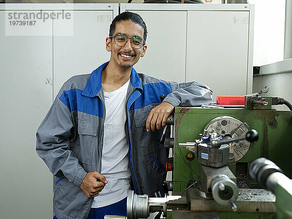 Smiling apprentice wearing eyeglasses leaning on railing at workshop