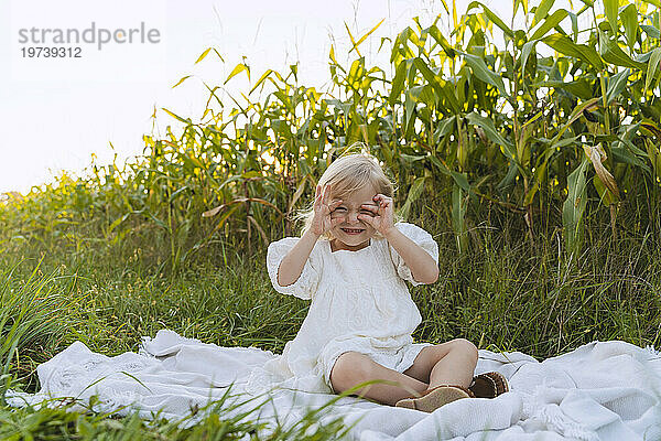 Glückliches süßes Mädchen gestikuliert und sitzt auf einer Decke vor Pflanzen
