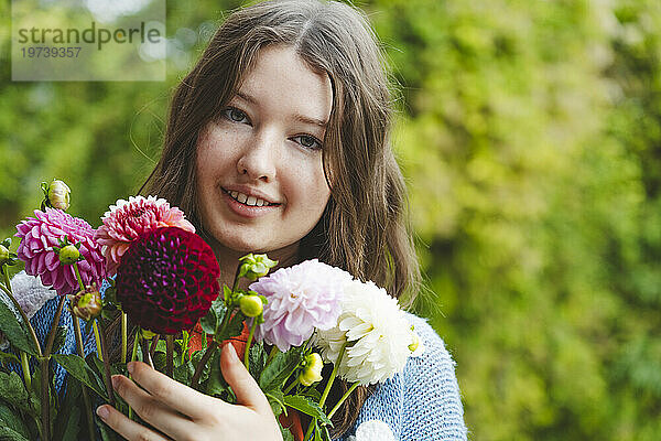 Lächelndes Teenager-Mädchen hält Dahlienblüten vor Pflanzen