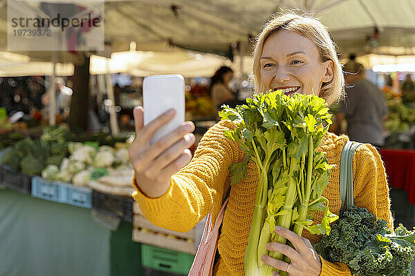 Frau macht mit Smartphone ein Selfie mit Sellerie auf dem Bauernmarkt