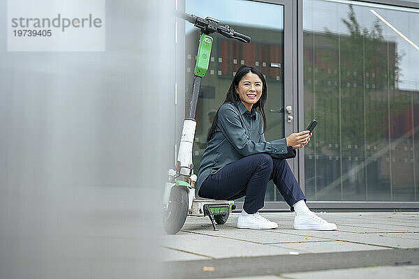 Glückliche Geschäftsfrau sitzt auf Elektroroller und Smartphone am Fußweg
