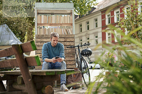 Reifer Mann benutzt Smartphone und sitzt neben Bücherschrank in einem Vorort