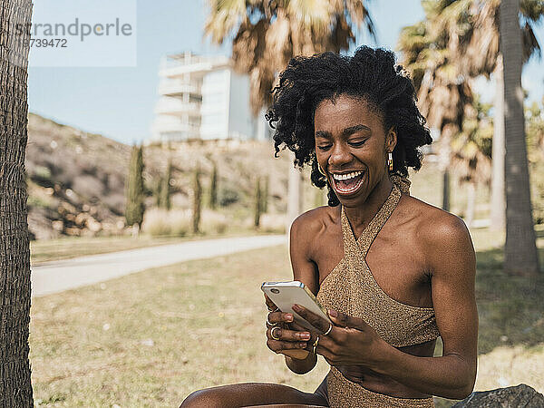 Fröhliche junge Frau im Bikini lacht und benutzt Smartphone im Park