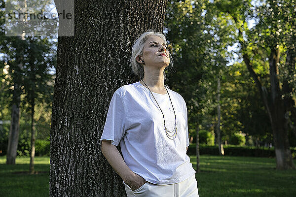 Reife Frau mit der Hand in der Tasche lehnt an Baum im Park