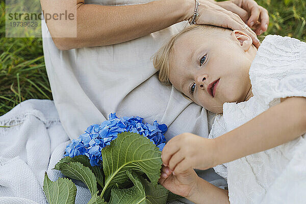 Blondes Mädchen mit blauen Blumen liegt auf dem Schoß der Mutter