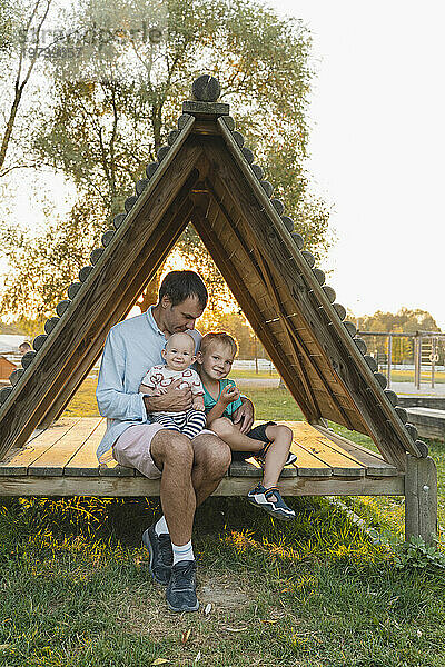 Liebevoller Vater sitzt mit Kindern in einer dreieckigen Holzhütte im Park