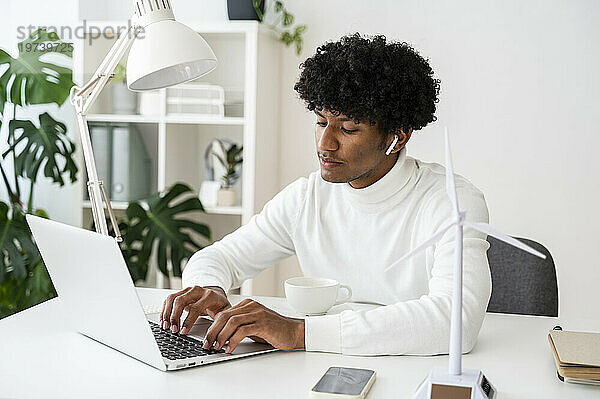 Konzentrierter Geschäftsmann mit Modell einer Windkraftanlage  der am Laptop am Schreibtisch im Heimbüro arbeitet