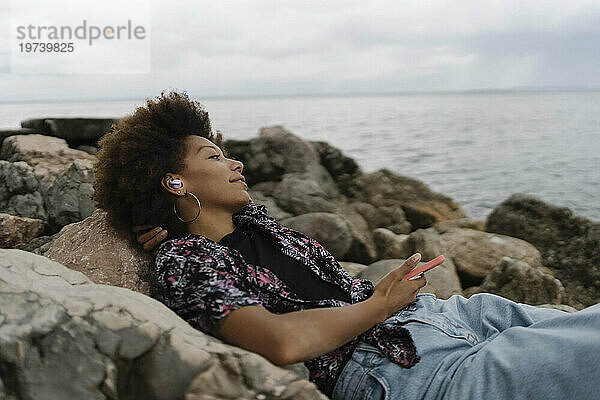 Lächelnde junge Frau entspannt sich auf Felsen und hört Musik über kabellose In-Ear-Kopfhörer