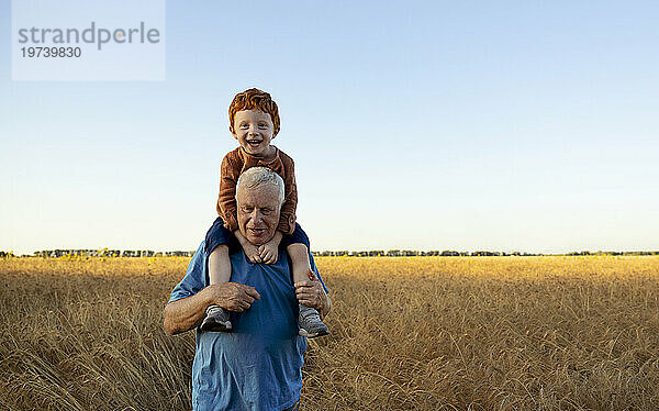 Glücklicher Großvater trägt Enkel auf Schultern im Weizenfeld unter Himmel