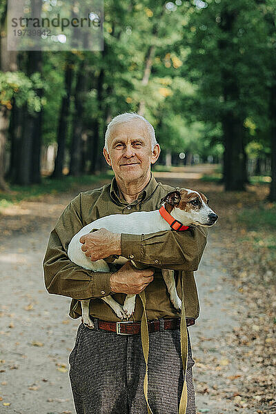 Smiling senior man holding Jack Russell Terrier dog in arms at park
