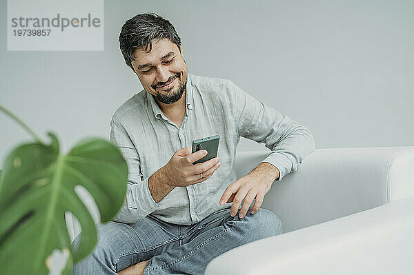Glücklicher Mann benutzt Smartphone auf Sofa vor weißer Wand